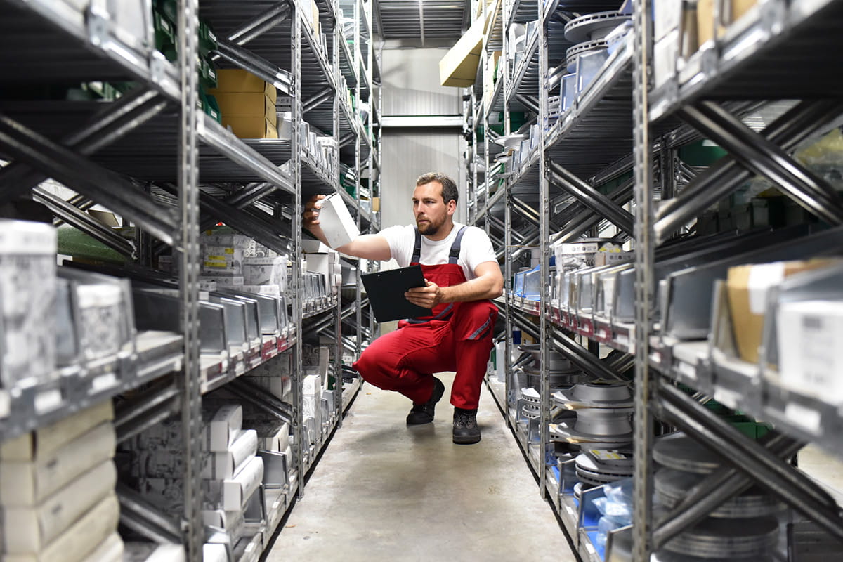 Zu sehen ist eine Person im Lager zwischen hohen Regalreihen, mit einem Tablet in der linken Hand und einem Päckchen in der rechten Hand.